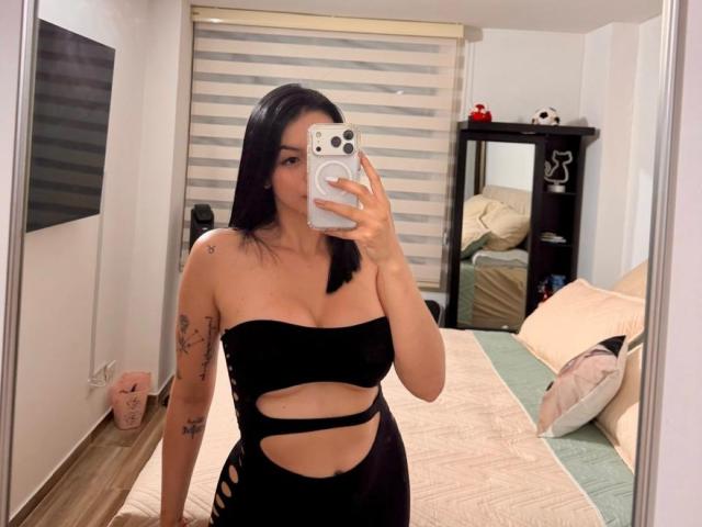IsabellaJames with black hair, wearing a black strapless cutout dress, showing her tits and ass, taking a mirror selfie, holding a white phone, visible tattoos on her arm.