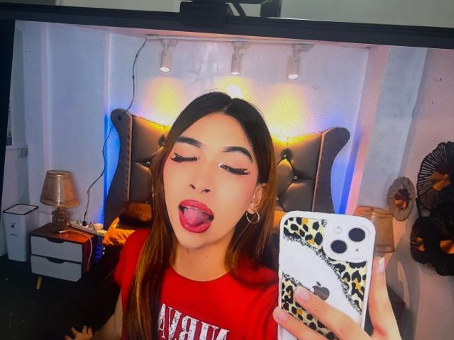 Magesticcock-atractivo, con pelo largo y castaño, camisa roja, lengua afuera, sosteniendo un teléfono con una funda de estampado de leopardo, en un dormitorio con cabecera negra acolchada y lámparas..