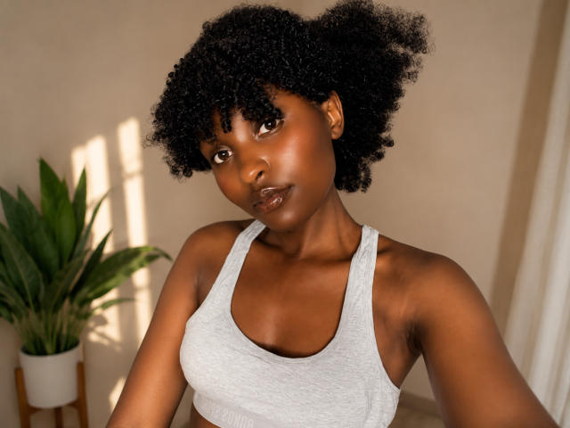 SpicyMelanin, con la pelle scura, i capelli neri ricci, indossa un reggiseno sportivo grigio, mostrando i suoi piccoli seni, e ha un'espressione neutra, mentre è in piedi in una stanza illuminata dal sole..