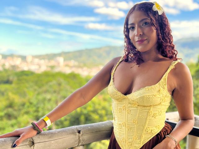 Mia Bianchi, con i capelli rossi ricci, un corsetto giallo con lacci e un fiore bianco, appoggiata a una ringhiera di bambù, che mostra il seno, indossa braccialetti, sorride alla telecamera, è una giornata di sole..