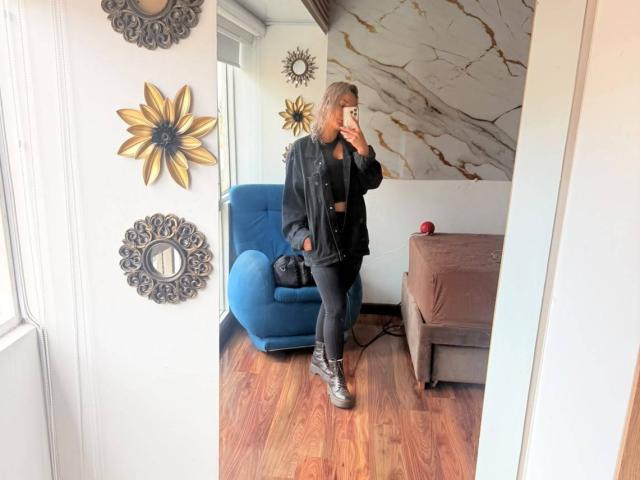 ElenaSaenz stands in a mirror, wearing a black jacket, black pants, and silver sneakers, holding her phone, with brown wooden floors and a blue chair behind her.