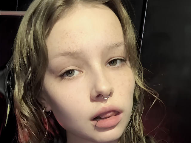 FletaDabbraccio a une peau pâle, des cheveux ondulés châtain clair, des yeux verts, un anneau au nez, un piercing aux lèvres, et de petites taches de rousseur, avec des lèvres légèrement entrouvertes et rosées..
