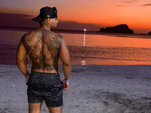 AdrianMontrell staat op een strand, met de rug naar de camera, en draagt een zwarte pet, een getatoeëerde borst en donkere, patroonrijke shorts. Hij kijkt naar een zonsondergang boven het water..