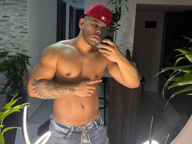 AdrianMontrell, sem camisa, usando boné vermelho, tatuagens no braço direito, segurando um telefone, olhando para o espelho, jeans, musculoso, pele morena, plantas verdes, luz de anel, fundo escuro, em pé, expressão séria..