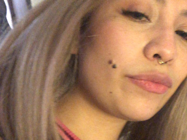 Ilaynna's face with blonde hair, pink lipstick, nose ring, and black moles on her cheek, looking slightly down. Her ass is not visible, only her face.