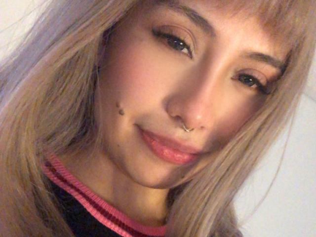 Ilaynna has light brown hair, a nose ring, and a small beauty mark on her cheek. She's smiling with pink lipstick and black eyeliner. Her tits are small and cute.