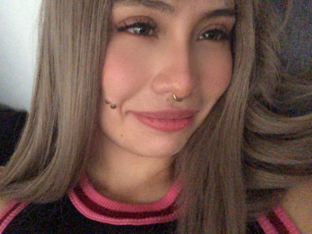 Ilaynna has long blonde hair, a nose ring, and a beauty mark on her chin. She's smiling with pink lipstick on, wearing a black top with pink stripes.