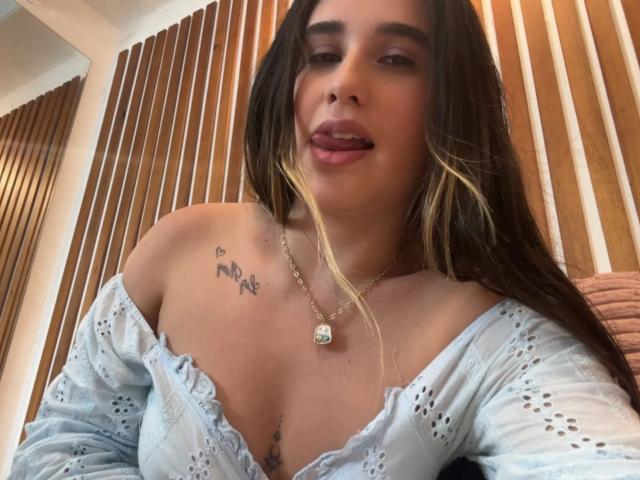 KatieDesirX con lunghi capelli castani, top leggero blu chiaro con una spalla scoperta, lingua fuori, piccolo tatuaggio sulla spalla sinistra, collana, che mostra il seno, pelle chiara e un'espressione carina e rilassata..
