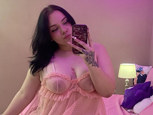 AlayaWillow with dark hair, pink sheer babydoll, showing her big boobs and tattooed hand, taking a mirror selfie in a pink-lit room, holding a phone with a floral case.