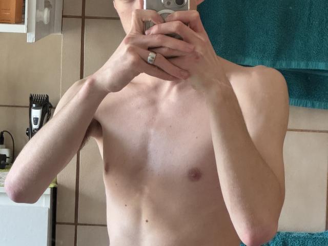 IceRagottiX sans chemise, avec un torse mince et pâle, de petits mamelons, tenant un appareil photo, portant une bague à la main droite, une serviette bleue en arrière-plan..
