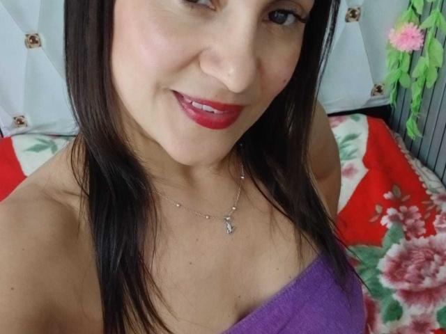 SolaraDubois, con cabello castaño oscuro, lápiz labial rojo, top morado sin tirantes, mostrando sus senos, un collar de plata pequeño, sonriendo, piel clara, en un selfie..