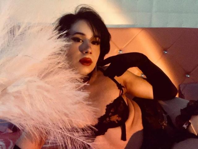 FemCupid con lingerie in pizzo nero, guanti neri e una piuma bianca, sdraiata su una testiera rosa trapuntata, che mostra i suoi seni e il rossetto rosso, guardando seducentemente la fotocamera..