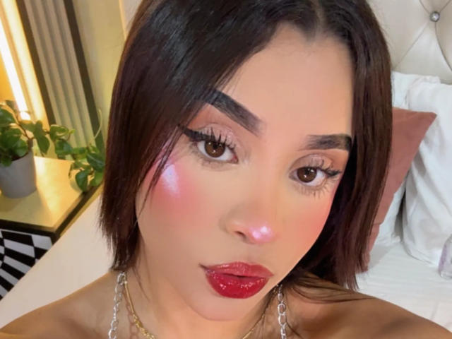 LunaVali a les lèvres rouges brillantes, les joues roses et luisantes, et les yeux marron foncé avec de longs cils. Elle porte un collier en chaîne et a les cheveux bruns et lisses..