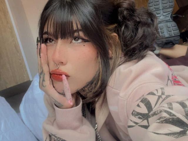 DirtyVenom, com cabelo escuro, franja e um piercing no nariz, sugando um brinquedo sexual rosa, vestindo um moletom rosa, com tatuagens na mão e no braço, sentada em um quarto..