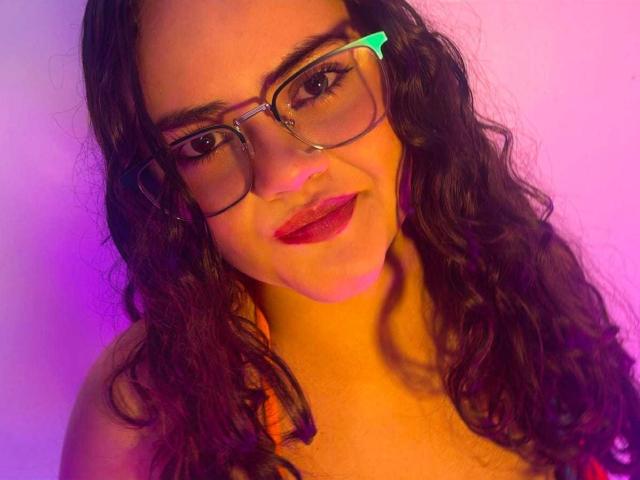 EmilyColsson has wavy brown hair, red lipstick, and wears glasses with a green clip. Her pink and purple lit face looks cute and slightly seductive.