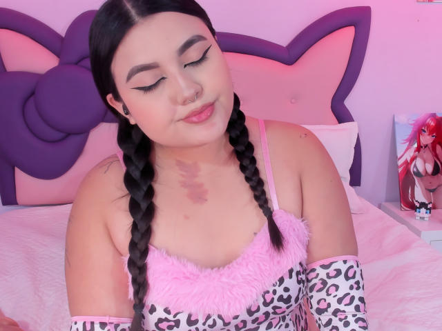 MarlaBriard, com cabelo preto trançado, delineador rosa, piercing no nariz e uma blusa rosa fofa, vestindo mangas com estampa de leopardo, sentada em uma cama rosa, olhando para baixo, fofa, sexy..
