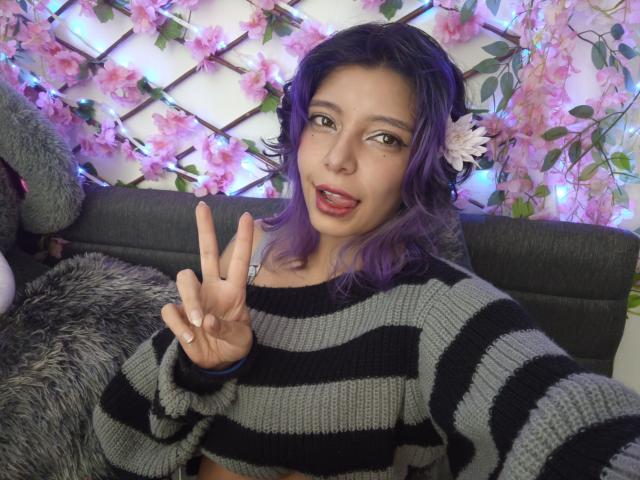 DarckVelvet con i capelli viola, un maglione a righe e un fiore tra i capelli, che mostra la lingua, fa un gesto di pace, ostenta il seno e il suo viso carino..