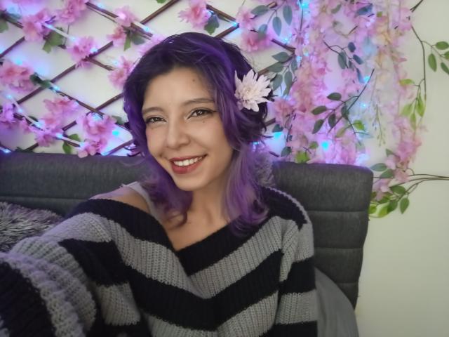 DarckVelvet mit lila Haaren, lächelnd, trägt einen grauen und schwarzen Streifenpullover, hat eine Blume im Haar, sitzt auf einem grauen Sofa und sieht mit blauen und pinken Lichterketten niedlich aus..