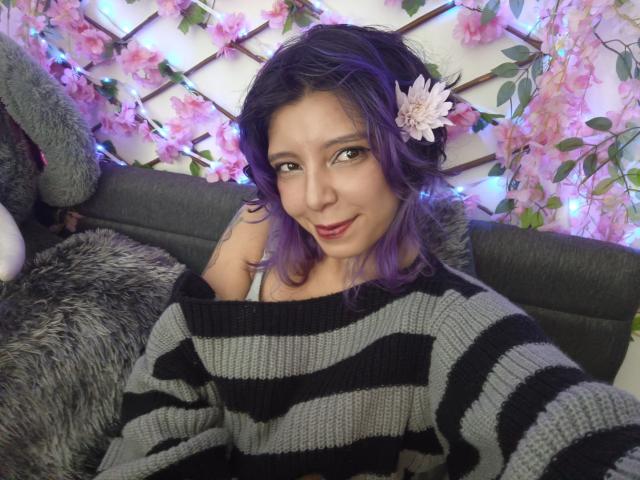 DarckVelvet, avec ses cheveux violets, une fleur dans les cheveux et un pull rayé, sourit à l'appareil photo, montrant légèrement ses seins dans le pull noir et gris..