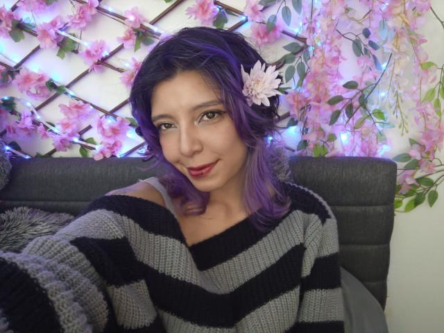 DarckVelvet mit lila Haaren, die einen gestreiften grauen und schwarzen Pullover trägt, hat eine rosa Blume im Haar, lächelt mit rotem Lippenstift und hat ein süßes, attraktives Gesicht..