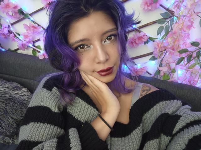 DarckVelvet, com cabelo roxo, suéter listrado preto e cinza, mão no rosto, batom vermelho, tatuagem no ombro esquerdo, sentada em um sofá cinza, cercada por flores rosa e luzes azuis..