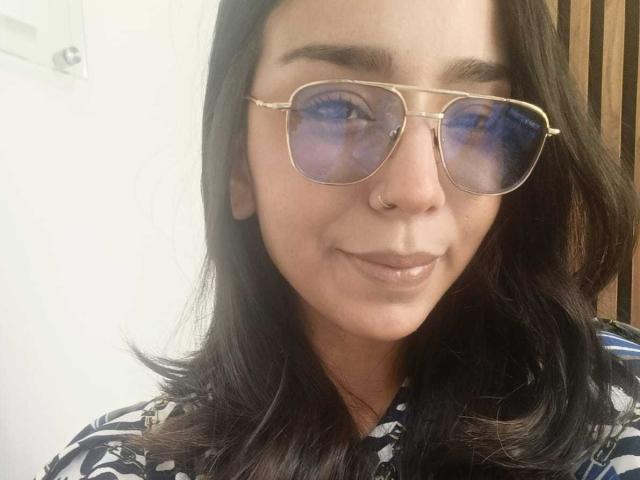 MilaThorne, con i capelli scuri, un anello al naso e occhiali da sole aviator dorati, sorride con le labbra lucide, indossando una camicia a righe bianche e nere..