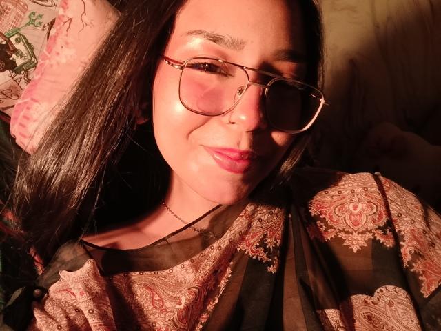 MilaThorne with long black hair, pink glasses, nose ring, and pink lipstick, wearing a sheer paisley top, smiling softly. Warm, glowing light on her face.