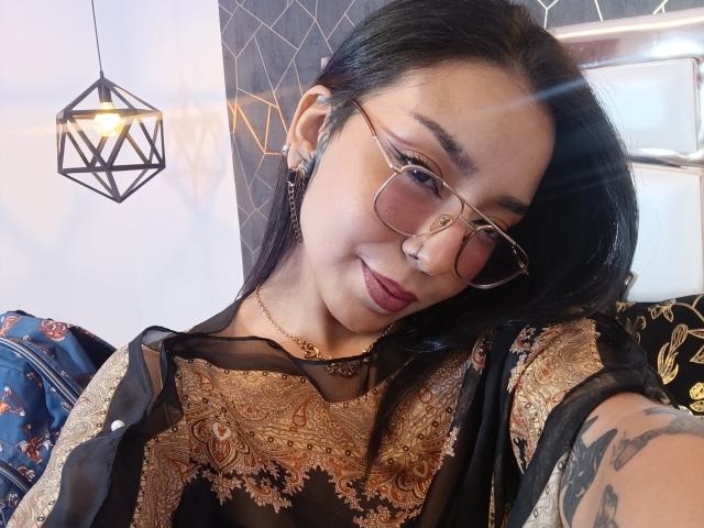 MilaThorne with dark hair, glasses, and red lipstick, wearing a black and gold patterned top, smiling at the camera, showing a tattoo on her right arm.