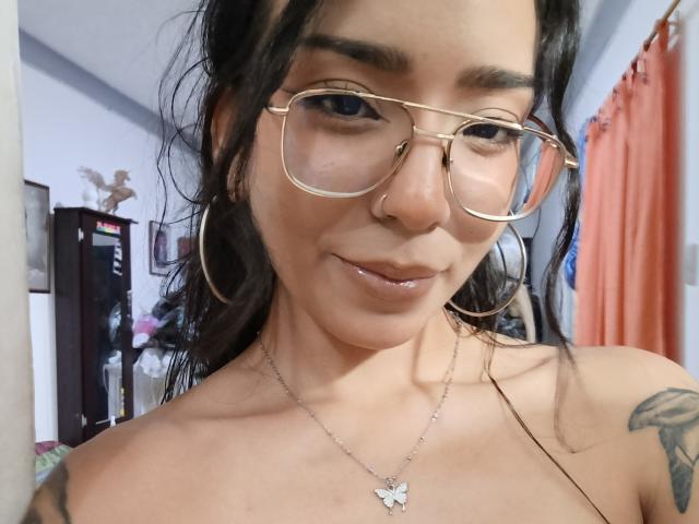 MilaThorne with glasses, nose ring, and butterfly necklace, has shiny lips, dark wavy hair, and a tattoo on her right shoulder, showing a subtle smile.