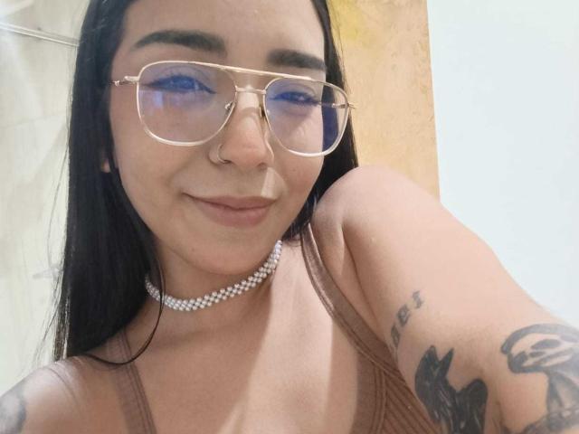 MilaThorne with long black hair, glasses, nose ring, choker, and tattoos, smiling at camera, wearing a brown tank top, showing her tits.