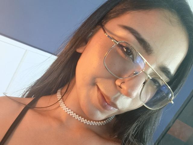 MilaThorne com óculos, colar de pérolas, piercing no nariz e blusa preta de alça fina, sorrindo para a câmera, cabelo castanho, pele clara, iluminação suave..