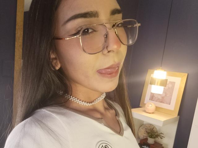 MilaThorne with long brown hair, wearing glasses, a pearl choker, and a white shirt, sticking out her tongue, showing her pink lips. Her ass is not visible.