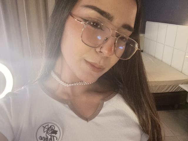 MilaThorne with long brown hair, wearing glasses and a pearl choker, has a cute face and small tits. She's in a white ribbed shirt with a logo.