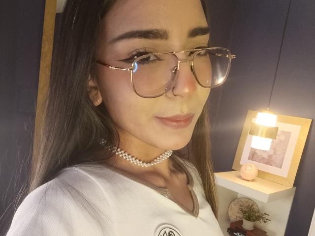 MilaThorne with long brown hair, wearing glasses, a pearl necklace, and a white shirt, showing a subtle smile, with soft lighting. Boobs not visible.