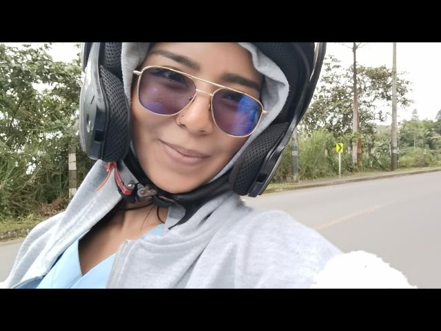 MilaThorne draagt een helm met oortjes, een blauw shirt, een grijze hoodie en reflecterende zonnebril, en glimlacht naar de camera met haar neusring. Op de achtergrond is een weg en groen te zien..