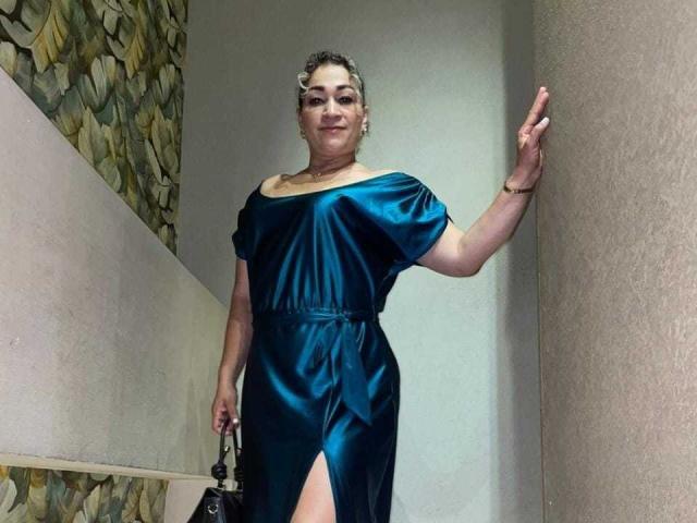 Samanttha Smith se tient dans un couloir, vêtue d'une robe turquoise brillante à une épaule, avec une fente haute, tenant un sac noir, souriant et touchant le mur avec sa main droite..