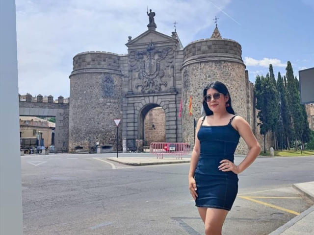 AmaraParker stands in a black, tight, sleeveless dress, showing her ass and tits, with hands on hips, in front of a stone, castle-like building, wearing sunglasses.