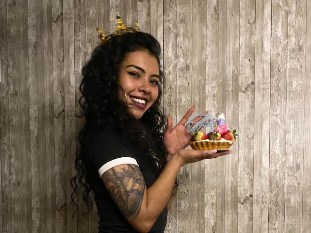 DevilAliceXo con capelli neri ricci, una corona d'oro, una maglietta nera, un braccio tatuato, che tiene un cono gelato con guarnizioni colorate, sorridente, con uno sfondo di parete in legno, in una foto casual e carina..