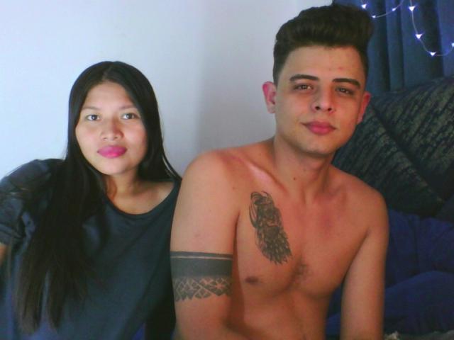 JenniDamian with long black hair, pink lipstick, wearing a dark shirt, sits next to shirtless JenniDamian with tattoos, dark hair, and a slight smile.