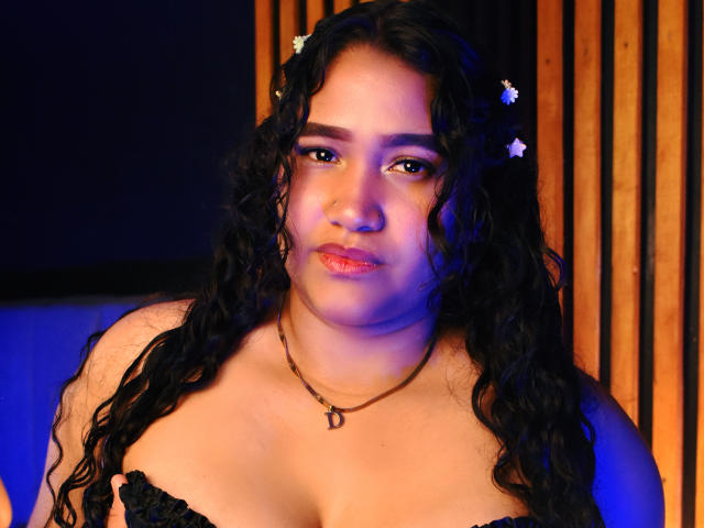 JoyceHill a les cheveux noirs bouclés avec des barrettes à fleurs, une poitrine de taille moyenne, des yeux marrons et un léger éclat bleu sur son visage. Elle porte une robe noire à bretelles et un collier en forme de "D"..