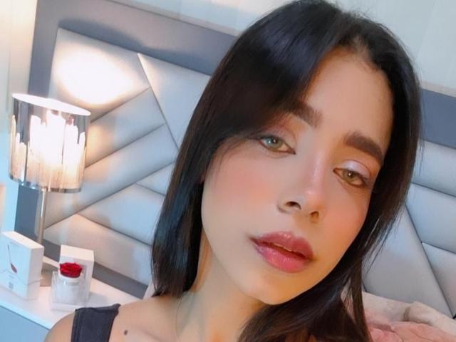 Samantha Stele hat glatte, braune Haut, langes, schwarzes Haar und glänzende, rosafarbene Lippen. Sie trägt ein schwarzes Oberteil, und links daneben befinden sich eine Lampe und eine Flasche mit rotem Verschluss..