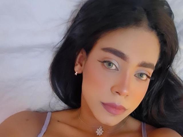 Samantha Stele heeft donker haar, groene ogen en roze lippenstift, draagt een ketting met een bloembedel en ligt op een wit kussen, met subtiele make-up en kleine oorbellen..