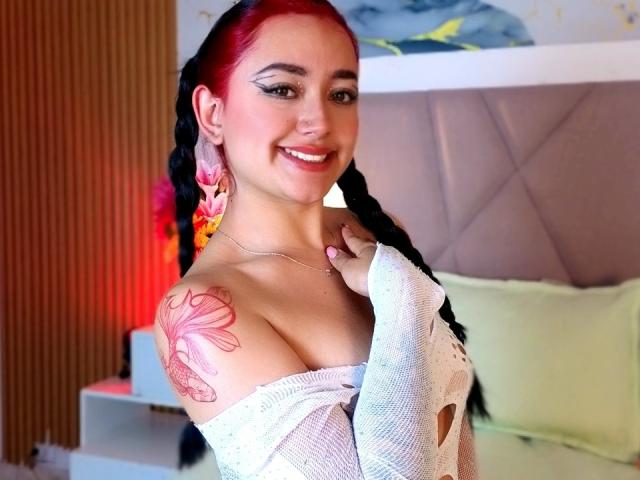 MiiaRousse, con trenzas rojas y negras, sonriendo y mostrando sus grandes senos con un tatuaje de una flor rosa en su hombro derecho, vestida con una blusa blanca con un hombro descubierto..