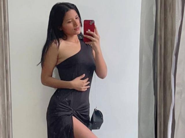 Daihana mit langen, schwarzen Haaren, die ein schwarzes Kleid mit einem Schulterträger trägt, ihre Hand auf den Bauch gelegt, mit einem hohen Schlitz am Bein, macht ein Selfie mit einem roten Handy..