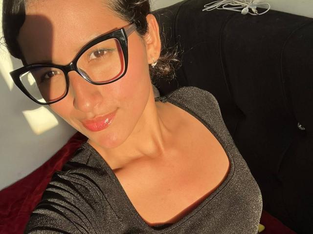 Daihana, com seios grandes, usando óculos pretos e uma blusa preta brilhante, tem um leve sorriso, lábios vermelhos brilhantes e está sob a luz do sol. Seus olhos castanhos estão fixos..