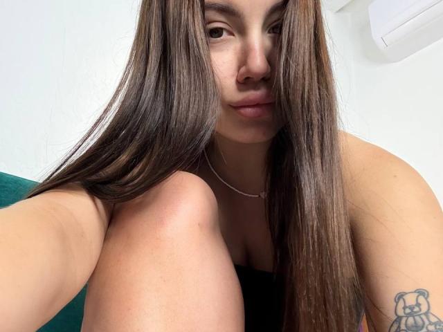 Ines Gueningsman, con lunghi capelli castani, labbra piene e un piccolo tatuaggio sul braccio destro, seduta con le ginocchia sollevate, indossa una maglietta nera che mette in mostra il seno..