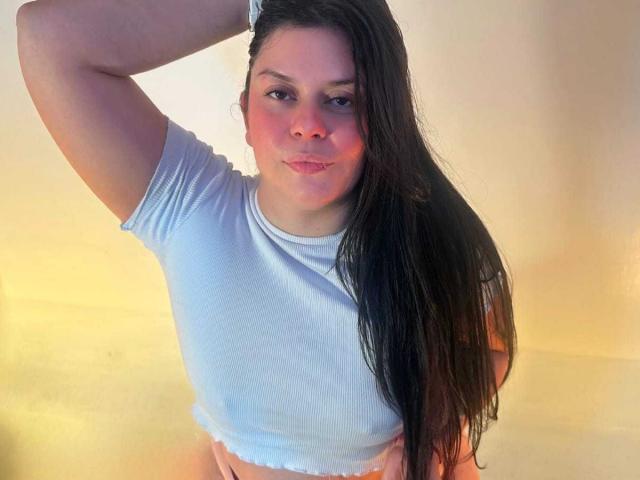 Charlotte Becker, con cabello largo y negro, vestida con una camisa azul claro, ligeramente rasgada, de manga corta, y con una pulsera en la muñeca, mirando a la cámara con una sonrisa leve y segura..