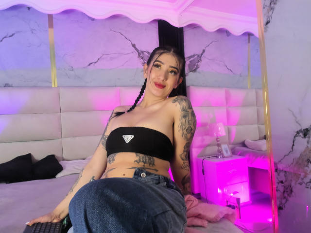 TifanyDiaz avec des tatouages, un haut noir sans bretelles laissant entrevoir de petites seins, des cheveux tressés, un jean bleu, assise sur un lit, avec des lumières roses et violettes, souriante, dans une chambre rose, avec une tête de lit blanche..