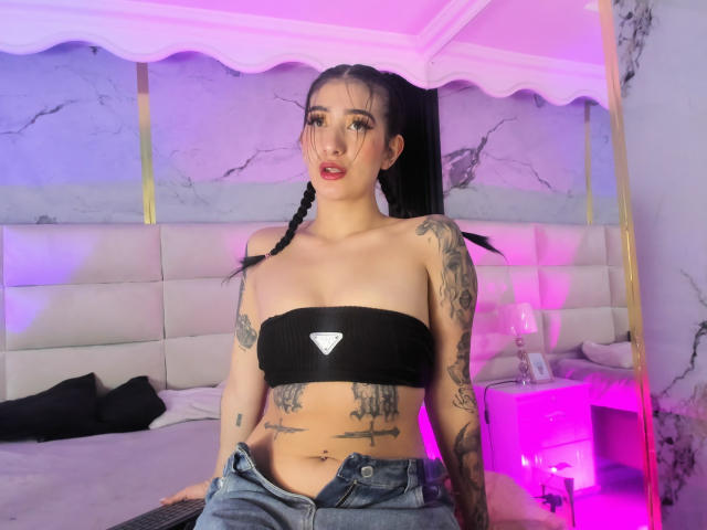 TifanyDiaz avec des tatouages, un haut noir sans bretelles laissant entrevoir de petites seins, un jean déboutonné, des cheveux tressés, une chambre rose et violette, des murs en marbre, assise sur le lit, regardant la caméra..