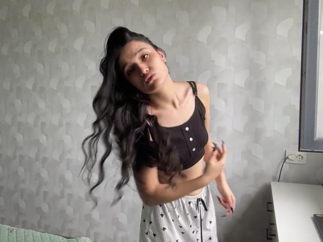 Youlanda Carlough, avec de longs cheveux noirs, portant un crop top noir et un pantalon de pyjama blanc, légèrement penchée en avant, regardant la caméra, la main droite près de sa poitrine..